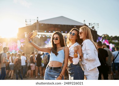 Group of happy young women friends take selfie at sunny beach music fest. Girls in trendy summer outfits enjoy live concert, party atmosphere. Youth culture, friendship, outdoor entertainment moment. - Powered by Shutterstock