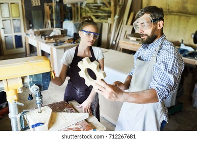 Group of hard-working carpenters looking at just finished wooden detail with concentration while standing at drill press machine, their aprons covered with sawdust - Powered by Shutterstock