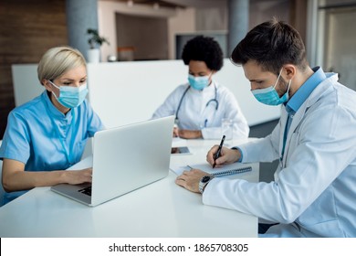 Group Of Healthcare Workers Wearing Face Masks While Working In The Office At The Hospital. Focus Is On Male Doctor Writing In Notebook.