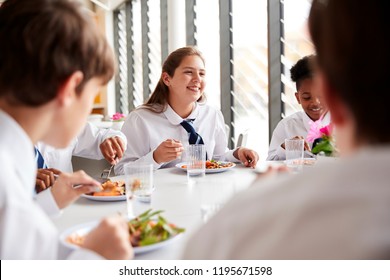 Group Of High School Students Wearing Uniform Sitting Around Table And Eating Lunch In Cafeteria