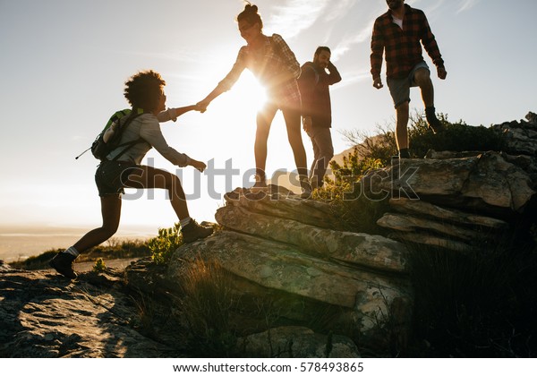 Group of hikers\
on a mountain. Woman helping her friend to climb a rock. Young\
people on mountain hike at\
sunset.