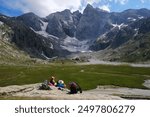 Group of hikers are sitting on rocks around Refuge des Oulettes de Gaube and looking to mountain massif with Vignemale - the highest peak of French Pyrenees. 