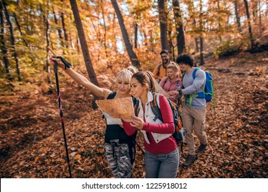 Group Of Hikers In Woods. Women Looking At Map And Leading The Rest Of The Group. Forest In Autumn Exterior.