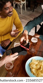Group of hispanic friends using NFC contactless technology with a smart phone in a bar. Customer paying the bill with mobile wireless bank payment terminal. 