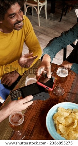 Group of hispanic friends using NFC contactless technology with a smart phone in a bar. Customer paying the bill with mobile wireless bank payment terminal. 