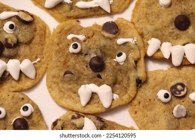 A group of homemade, imperfect, chocolate chip cookies, crowded onto a white plate.  Each has a face  of candy eyes and marshmallow teeth.  Some have marshmallow eyebrows and chocolate chip noses - Shutterstock ID 2254120595