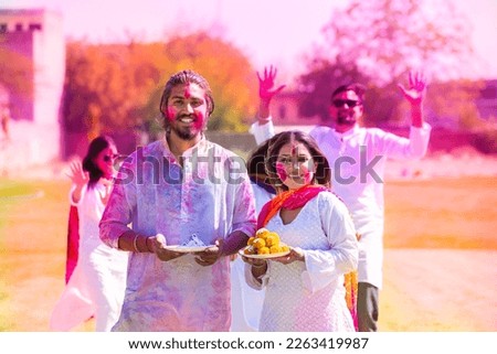 Group of indian friends wearing white kurta and holding plate full of laddu sweet and gula celebrating holi festival wtih colorful people at park outdoor.