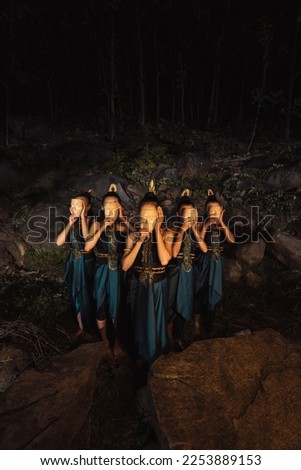 A Group of Indonesian women holding and wearing a brown wooden mask in a green skirt between the rock inside the woods