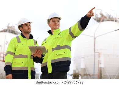 group of industrial engineers workers in a refinery - oil and gas processing equipment and machinery, engineers collaborate with a laptop, blueprint, and digital tablet at the oil storage tanks site.