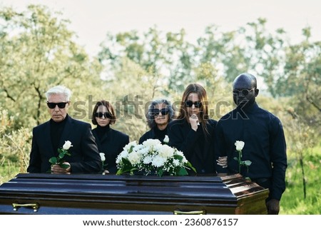 Group of intercultural grieving people in mourning attire and sunglasses holding white roses while standing in front of coffin with closed lid