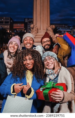 Group of international friends with shopping bags and a christmas gift smiling, laughing and looking at camera. Happy buddies celebrating the christmastime. Young adult people enjoying the xmas night
