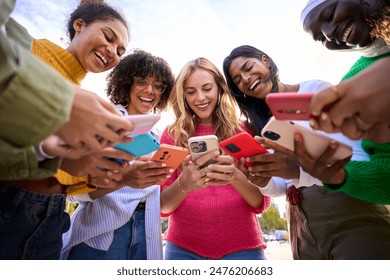 Group of international happy generation z only women laughing gathered in circle looking at cell phones outdoors. Cheerful young females using mobiles with funny expression. Addiction to technology 
