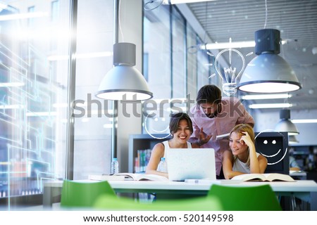 Group of international university students having fun studying in library, three colleagues of modern work co-working space talking and smiling while sitting at the desk table with laptop computer