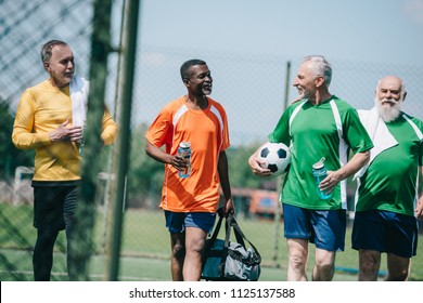 Group Of Interracial Elderly Sportsmen With Sportive Water Bottles Walking On Football Field