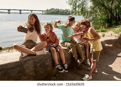 Group of joyful multiracial children taking selfie on smartphone on river coast outdoors. Boys and girls of generation alpha. Gadget addiction. Friendship. Childhood lifestyle. Sunny summer day - Powered by Shutterstock