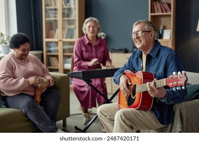 Group of joyful senior people playing music together enjoying talent show in retirement home focus on smiling senior man playing guitar copy space - Powered by Shutterstock