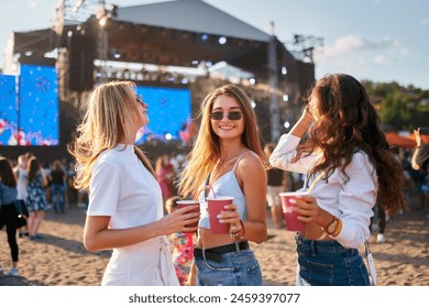 Group of joyful women with cool summery drinks at beach music festival. They dance under clear sky, stage with live performance in background. Girls in sunglasses enjoy fest vibes, sunlit ambiance. - Powered by Shutterstock