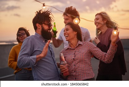 Group of joyful young friends in casual clothes drinking beer and dancing while enjoying summer party on rooftop at sunset time - Shutterstock ID 1845065842