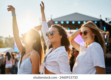 Group of joyful young women dance at sunny beach music festival. Girls in summer outfits enjoy concert, hands up in celebration. Vibrant fest atmosphere, friends share fun experience by sea. - Powered by Shutterstock