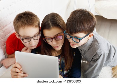 Group Of Kids In Eye Glasses Look Into Tablet. Children Computer Games, Social Networks And Media Addiction Concept. Girl And Boys With Tablet. Communication Technologies. Eyesight, Eyewear For Kids. 