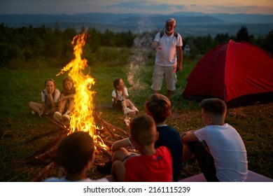 Group Of Kids Listening Senior Instructor, While Sitting Around Campfire In Mountains. Front View Of Bearded Guide Giving Instruction For Children, Telling Stories In Campsite. Concept Of Camping.