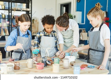 Group of kids and their teacher leaning over table with gouahes and painting self-made clay items - Powered by Shutterstock