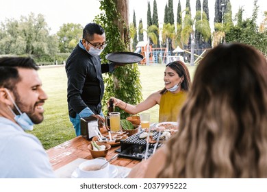 Group Of Latin Friends With Masks On The Terrace Of Mexican Restaurant Or Bar Attended By A Waiter With Face Mask. Social Distancing During Coronavirus Pandemic