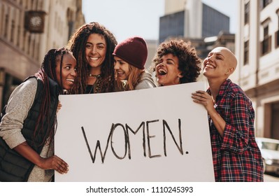 Group Of Laughing Demonstrators On Road With A Women Signboard. Female Activists Enjoying During A Protest For Women.