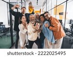 Group of laughing multiracial coworkers dressed in casual clothes laughing and looking at mobile phone screen while taking self shot and having fun during break in office