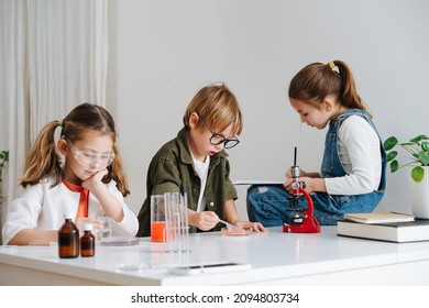 Group Little Kids Doing Home Science Project, Working With Chemical Reagents, Girl Is Documenting The Process. All Sitting Behind The Table.