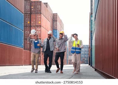Group of logistics workers walking at container terminal after finishing work, Diverse technical team in safety gear walking past cargo containers at port