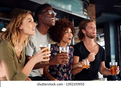 Group Of Male And Female Friends Celebrating Whilst Watching Game On Screen In Sports Bar