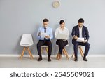 Group of male and female job seekers sitting in row on chairs waiting for job interview. Young people holding paper documents and resumes, using laptop, waiting their turn. Human resources, employment