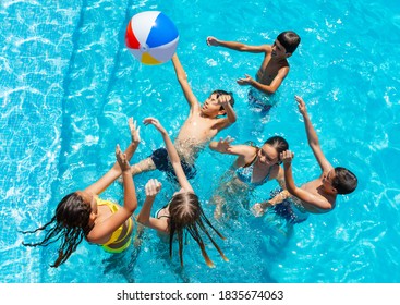 Group Of Many Kids Jump After Ball And Play In Swimming Pool, Splashing, Lift Hands Smile View From Above