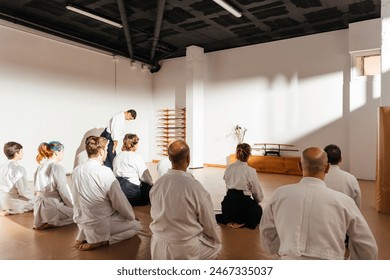 A group of martial arts students, dressed in white gi, attentively sit in the traditional seiza position as an instructor oversees the practice in a sunlit dojo. - Powered by Shutterstock