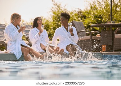 Group Of Mature Female Friends Wearing Robes Sitting With Feet In Pool Drinking Champagne On Spa Day - Powered by Shutterstock