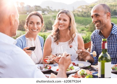 Group Of Mature People Eating Together In A Vineyard In A Summer Day. Happy Woman Sipping Wine While Talking To Friends During A Lunch In A Winery. Senior Couple Having Dinner With Wine At Sunset.