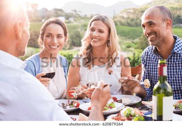 Group of mature people eating together in a\
vineyard in a summer day. Happy woman sipping wine while talking to\
friends during a lunch in a winery. Senior couple having dinner\
with wine at sunset.