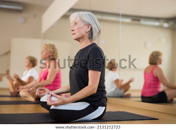 Group of mature women doing yoga in a\
fitness studio meditates in a half-lotus\
position