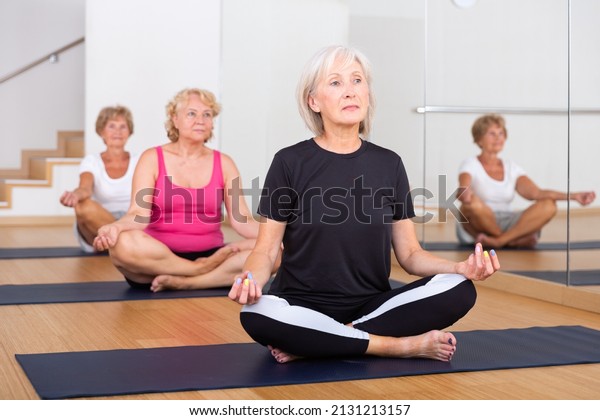 Group of mature women doing yoga in a\
fitness studio meditates in a half-lotus\
position