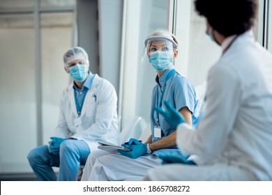 Group of medical experts wearing face shields and masks while communicating in a hallway and sitting apart from each other due to coronavirus pandemic. Focus is on nurse. 