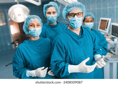 Group of medical professionals standing and looking at camera in operating room as they pose together for a portrait, dressed professionally in operating gowns, wearing surgical masks, caps and gloves - Powered by Shutterstock