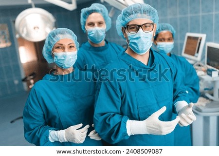 Group of medical professionals standing and looking at camera in operating room as they pose together for a portrait, dressed professionally in operating gowns, wearing surgical masks, caps and gloves