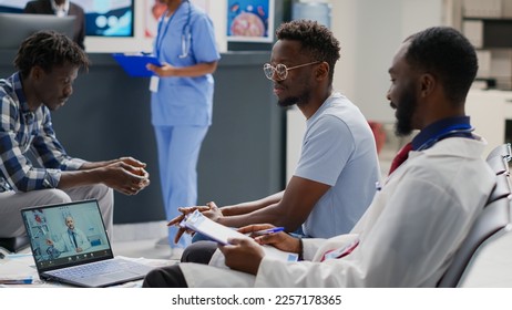 Group of men meeting with physician on remote videocall, using webcam on laptop in waiting area. Doctor and patient sitting in hospital reception to talk to medic on online telemedicine call. - Powered by Shutterstock