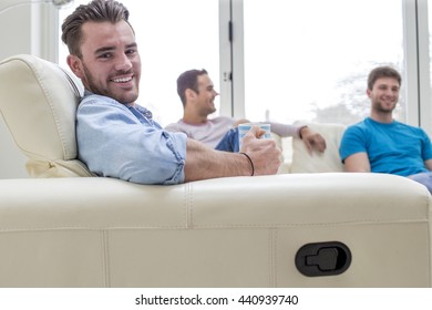 A Group Of Men Are Sat At Home Talking And Laughing. One Man Is Looking At The Camera Smiling.