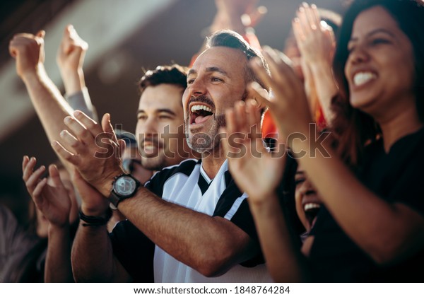 Group of men and women cheering their national\
team. Football team supporters enjoying during watching a live\
match from stadium.