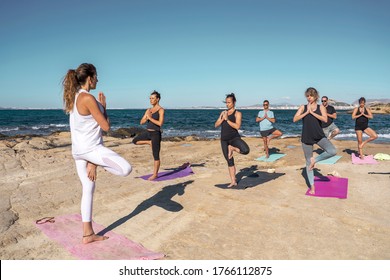 Group of men and women doing yoga on a beach on sunny day by the sea. The group is led by a yoga teacher dressed in white sportswear. - Powered by Shutterstock