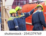 A group of merchant ship crew members stands together during an abandon ship drill, discussing the procedure for lowering the lifeboat. Their coordinated focus highlights the importance of teamwork.