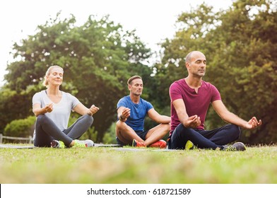 Group Of Middle Aged People Doing Yoga Sitting On Grass. Three People Practicing Meditation And Yoga At Park On A Bright Morning. Mature Woman And Two Mid Men Meditating Together In A Lotus Position.