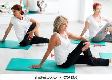 Group Of Middle Aged Women Warming Up In Gym.Threesome Sitting On Floor On Rubber Mattresses.